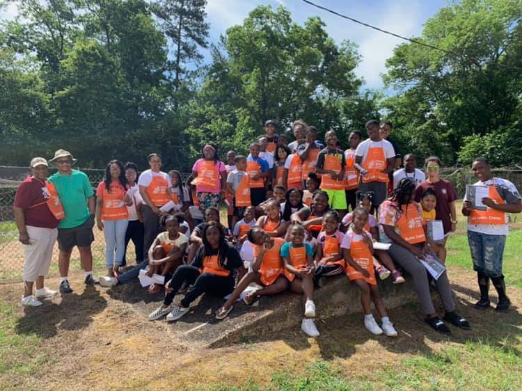 A group of  several African American children sitting on a dirt pile wearing orange volunteer vests.